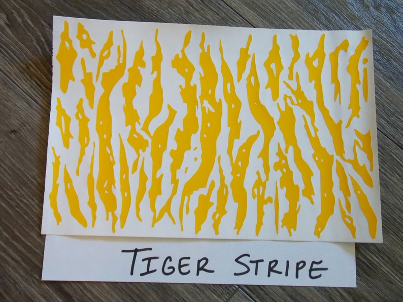 K&ouml;nnte beinhalten: Eine Folie mit gelbem Tigerstreifen-Camouflage. Die Folie ist mit einem sich wiederholenden Muster aus gelben Streifen auf wei&szlig;em Hintergrund bedruckt. Der Text "TIGER STRIPE" ist in Schwarz auf wei&szlig;em Hintergrund unter der Folie geschrieben.