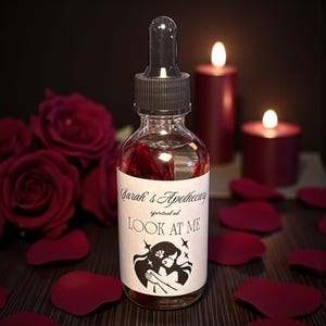 Puede incluir: Una botella de vidrio transparente de aceite espiritual con un cuentagotas negro, etiquetada como "Sarah's Apothecary" y "LOOK AT ME". La etiqueta presenta una silueta de dos figuras. Pétalos de rosa rojos y velas encendidas rodean la botella.