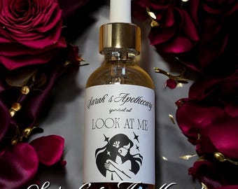 May include: A clear glass dropper bottle with a gold collar and white cap. The label features the text Sarah's Apothecary, spiritual oil, and LOOK AT ME, along with a black graphic illustration of a person with long hair and stars.