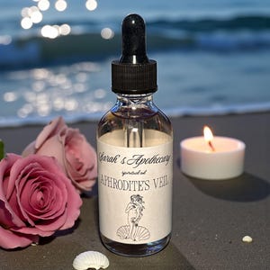 May include: A glass bottle of Aphrodite's Veil spiritual oil with a black dropper, on a sandy surface. The label has text and a line drawing of a woman. A lit candle and pink roses are also visible in the image.