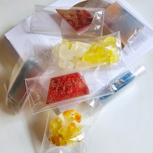 May include: An assortment of colorful, individually packaged candies and treats. The image features various shapes and colors, including red, yellow, and white, presented in clear plastic bags. The treats are arranged on a white surface, suggesting a gift or sample set.