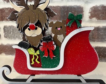 May include: A wooden Christmas decoration featuring a Highland cow dressed as a reindeer pulling a red sleigh with a teddy bear and presents. The sleigh is decorated with glitter and snowflakes.