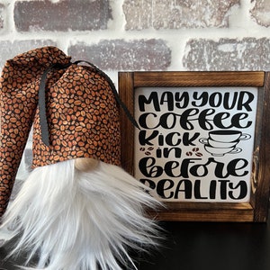 Puede incluir: Un gnomo blanco con un gorro de patrón de granos de café marrón y negro y una barba blanca. El gnomo está de pie frente a un letrero de madera que dice "Que tu café te dé un empujón antes de la realidad".
