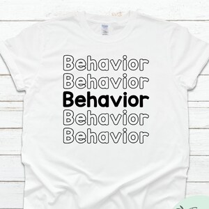 May include: White t-shirt featuring the word "Behaviour" in a repeating pattern. The word "Behaviour" is in black, outlined in white, with one instance in solid black. The Anne Marie Apparel logo is in the bottom right corner.