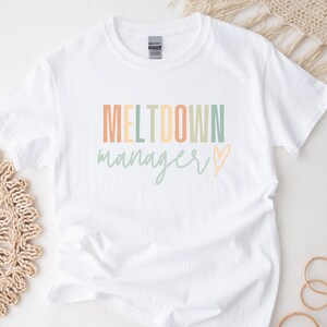 Puede incluir: Camiseta blanca con el texto "MELTDOWN manager" en colores arcoíris con un corazón al final.