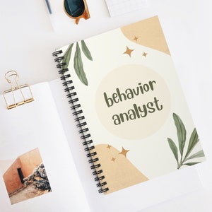 May include: A white spiral bound notebook with a beige and green floral design. The notebook has the text "behavior analyst" in brown lettering.