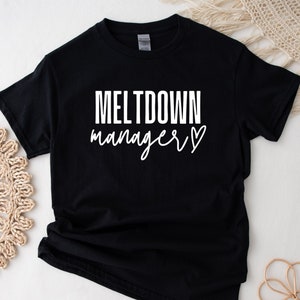 Puede incluir: Camiseta negra con texto blanco que dice "MELTDOWN manager" con un pequeño símbolo de corazón.