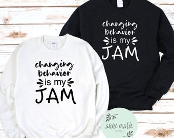 May include: Two white and black sweatshirts with the text "changing behavior is my JAM" printed on them. The sweatshirts are laid out on a wooden surface.