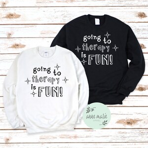 Puede incluir: Dos sudaderas con el texto "going to therapy is FUN!" impreso en ellas. Una sudadera es blanca y la otra es negra.