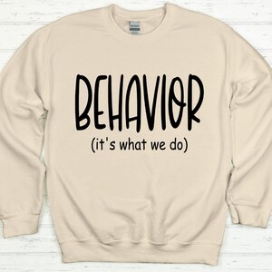 以下が含まれることがあります： ベージュのトレーナーに、黒い文字で「BEHAVIOR (it's what we do)」と書かれています。