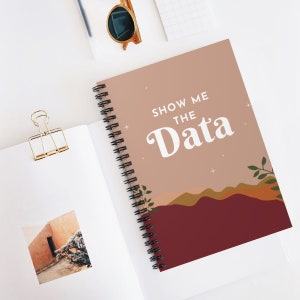 以下が含まれることがあります： 茶色の表紙に「Show Me The Data」という白い文字が書かれた、スパイラルバインディングのノート。背景は、山と葉のついた枝のある夕焼けの風景です。