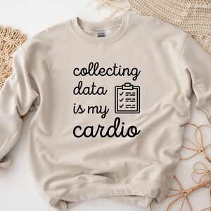Könnte beinhalten: Ein beiger Pullover mit dem Text "collecting data is my cardio" und einem schwarz-weißen Checklisten-Symbol.
