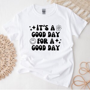 May include: White t-shirt with black text that reads "It's a good day for a good day". The text is in a retro font style and includes a smiley face and a flower graphic.