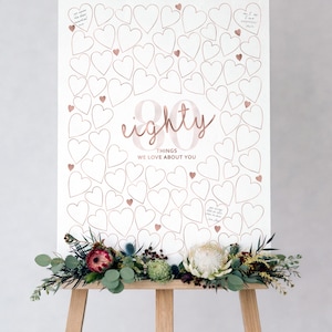 May include: A white rectangular sign on a wooden easel featuring rose gold outlines of numerous hearts. The center displays the text "80" in large rose gold font, with "THINGS WE LOVE ABOUT YOU" printed below. Floral arrangements decorate the base.