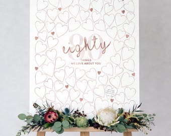 Puede incluir: Un póster blanco sobre un caballete de madera con contornos de corazones en oro rosa y el texto 80 THINGS WE LOVE ABOUT YOU. El póster está decorado en la base con un arreglo floral que incluye una protea blanca, una flor roja y hojas de eucalipto.