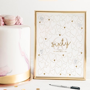 May include: A gold-framed sign featuring the text 60 THINGS WE LOVE ABOUT YOU surrounded by numerous outlined hearts, accompanied by a white cake topped with pink macarons and a black marker pen for writing messages on the sign.