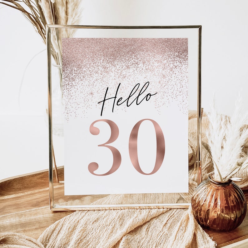 May include: A framed sign with the words "Hello 30" in black script and rose gold numbers. The top of the sign has a rose gold glitter effect. The sign is displayed in a clear glass frame, with decorative elements in the background.