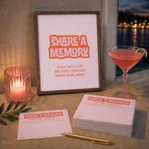 Op de afbeelding: Een ingelijst bord met oranje tekst SHARE A MEMORY, PLEASE TAKE A CARD AND SHARE A TREASURED MEMORY OR WELL WISHES, vergezeld van een stapel bijpassende kaarten, een enkele kaart, een gouden pen, een roze kaars in glas en een cocktail.