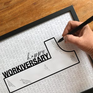 以下が含まれることがあります： 「Happy Workiversary」の文字が黒い太字で書かれた額入りのプリント。背景には「1年間の勤務おめでとうございます！」というテキストが繰り返されています。手はペンを持っており、プリントに書き込む準備ができています。