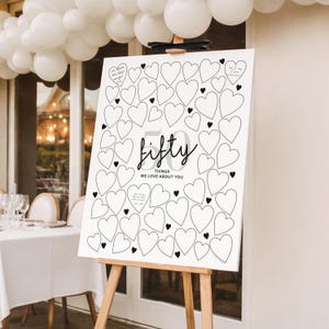 Peut inclure: Un panneau blanc sur un chevalet en bois, avec un motif de c&oelig;urs et les mots "fifty things we love about you." Le panneau est d&eacute;cor&eacute; de c&oelig;urs et de texte noirs, parfait pour un mariage ou un anniversaire.