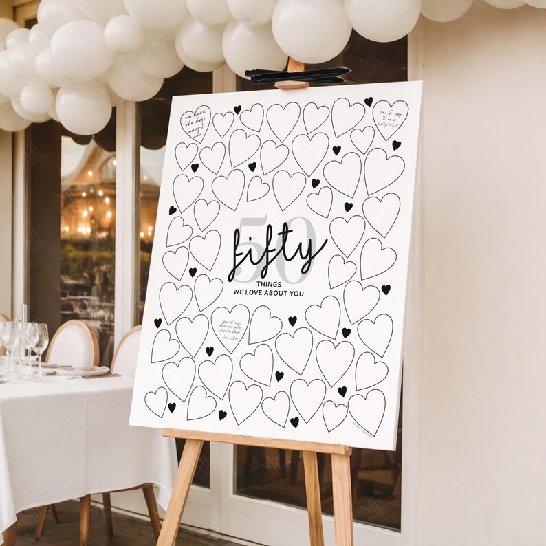 Peut inclure: Un panneau blanc sur un chevalet en bois, avec un motif de c&oelig;urs et les mots "fifty things we love about you." Le panneau est d&eacute;cor&eacute; de c&oelig;urs et de texte noirs, parfait pour un mariage ou un anniversaire.