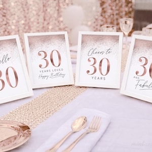 May include: Four framed signs with the number 30 in rose gold, with phrases like "Hello 30" and "Cheers to 30". The signs are on a table with gold cutlery and a decorative runner, perfect for a birthday celebration.