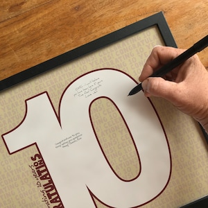 Può includere: Una stampa incorniciata con un grande numero 10 bianco, bordato di bordeaux, con le parole "Congratulazioni" e "Festeggiando 10 anni" in un motivo ripetuto. Una mano sta scrivendo sul numero 10 con una penna nera.