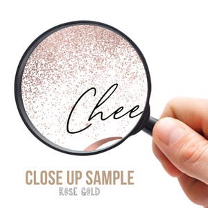 May include: A close-up view through a magnifying glass reveals the word "Chee" in elegant black script, set against a backdrop of rose gold glitter. The words "CLOSE UP SAMPLE ROSE GOLD" are below the magnifying glass.