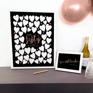 May include: A white framed sign with a black background and white hearts. The sign says "50 things we love about you" in rose gold lettering. A smaller white framed sign says "guestbook please sign" in rose gold lettering.