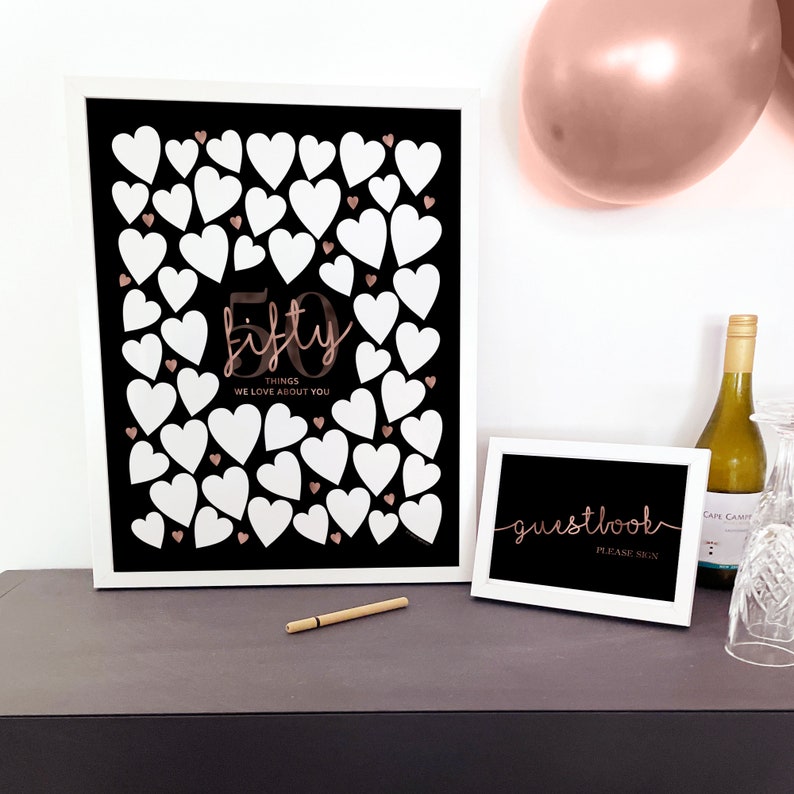 May include: A white framed sign with a black background and white hearts. The sign says "50 things we love about you" in rose gold lettering. A smaller white framed sign says "guestbook please sign" in rose gold lettering.