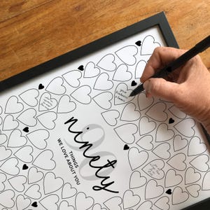 May include: A framed print with a black border featuring a design of hearts. The print includes the text "ninety things we love about you". A hand is writing on the print with a black pen, filling in the heart shapes with personalized messages.