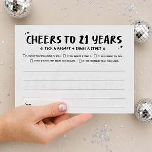 以下が含まれることがあります： CHEERS TO 21 YEARSおよびTICK A PROMPT + SHARE A STORYという黒い文字が印刷された白い長方形のカード。5つのチェックボックスと記入用の4本の点線があり、小さな星のアイコンで装飾されています。