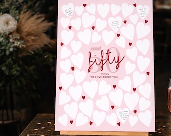 May include: A pink canvas sign on a wooden easel, featuring white heart shapes and small red hearts. The sign reads "fifty things we love about you" in red script. Small handwritten notes are scattered across the hearts.