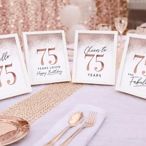 May include: Four framed signs with the number 75 in rose gold. The signs say "Hello 75", "75 Years Loved Happy Birthday", "Cheers to 75 Years", and "75 and Fabulous". The signs are on a table with gold accents.