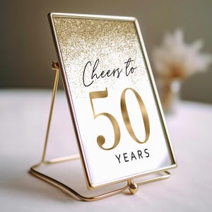 Gold glitter table number card with a gold frame and stand. The poster says "Cheers to 50 Years".