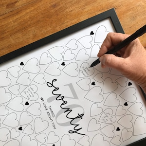 May include: A framed print with a black border featuring a design of numerous heart shapes. The words "seventy things we love about you" are displayed in a stylish font. A hand is writing on one of the hearts with a black pen.