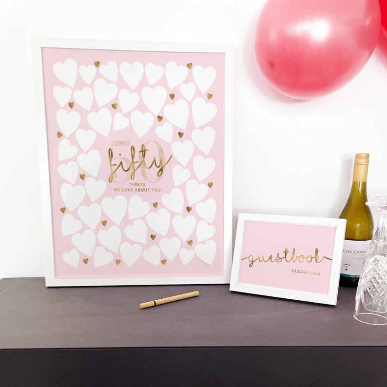 May include: A white framed sign with a pink background and gold lettering that says "fifty things we love about you". The sign is decorated with white and gold hearts. A smaller white framed sign with a pink background and gold lettering that says "guestbook please sign" is also visible.
