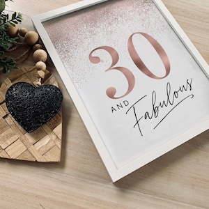 May include: A framed print with the text "30 and Fabulous" in rose gold and black script. The background has a rose gold glitter effect. The frame is white. A black heart-shaped decoration and wooden beads are in the foreground.