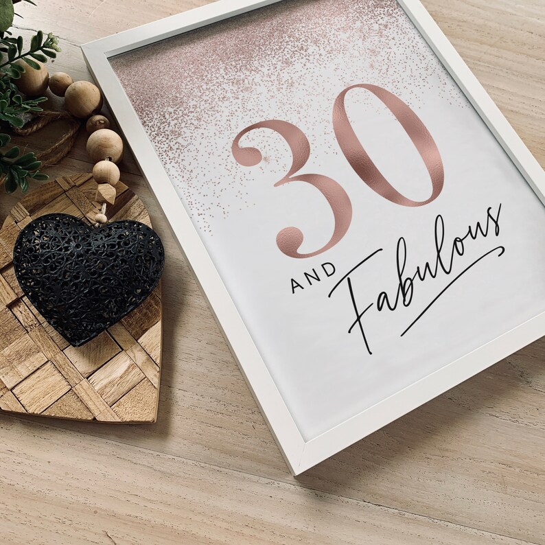 May include: A framed print with the text "30 and Fabulous" in rose gold and black script. The background has a rose gold glitter effect. The frame is white. A black heart-shaped decoration and wooden beads are in the foreground.