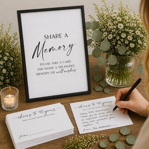 May include: A black framed sign with the words "SHARE A Memory" in elegant script. Below, text reads "PLEASE TAKE A CARD AND SHARE A TREASURED MEMORY OR well wishes." Beside the sign are cards and a pen. A bouquet of flowers is in the background.