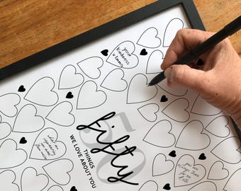 May include: A framed print with a black border and a design of hearts. The text reads "Fifty Things We Love About You". A hand is writing on the print with a black pen, adding personalized messages within the heart shapes.
