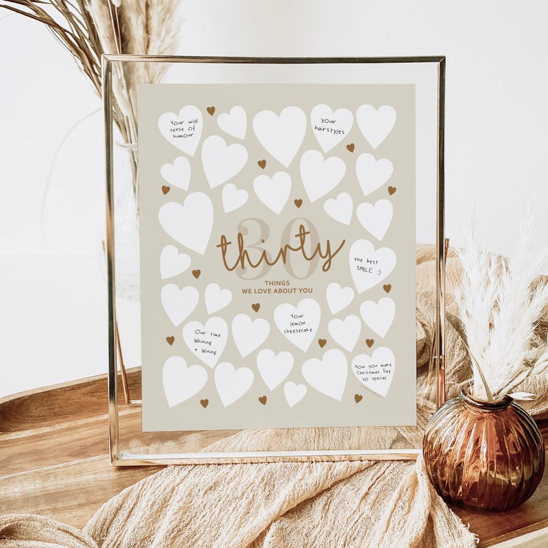 30th birthday sign with "Thirty Things We Love About You" writing, surrounded by 30 blank hearts, on beige background, in gold floating frame.
