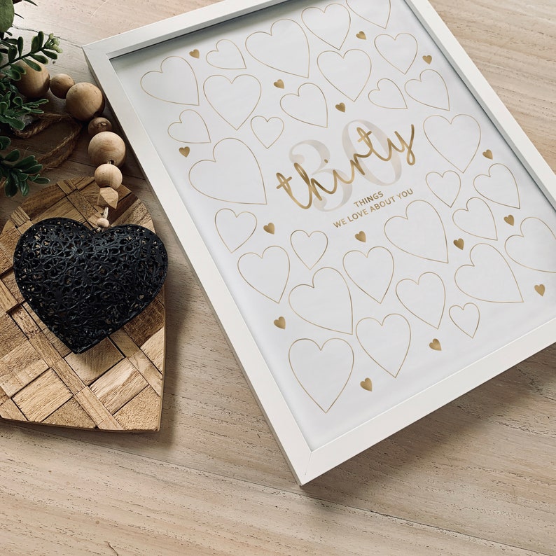 Puede incluir: Impresi&oacute;n enmarcada en blanco con una inscripci&oacute;n dorada que dice "treinta cosas que amamos de ti". La impresi&oacute;n presenta un fondo blanco con un patr&oacute;n de corazones dorados.