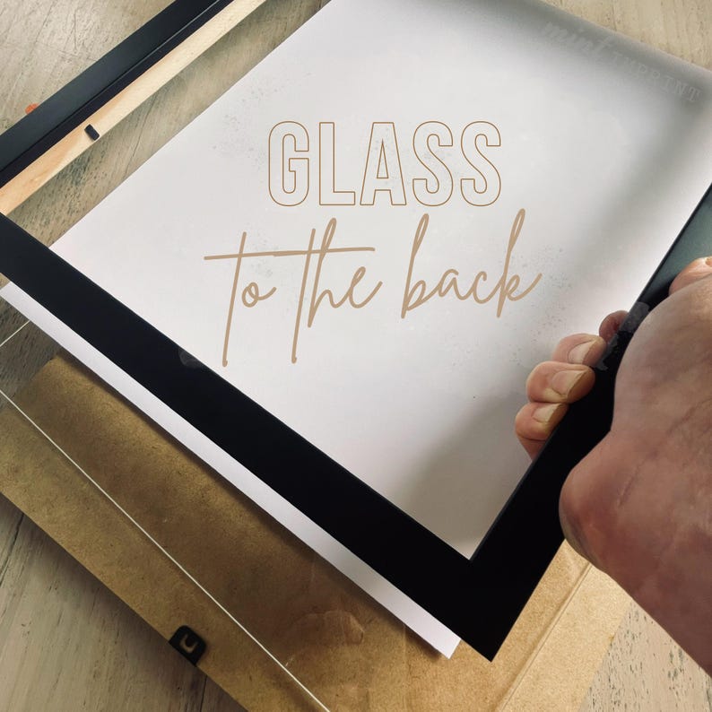 May include: A black picture frame with the text "GLASS to the back" in a light brown color. The frame is being held by a person. The frame is on top of a white paper and a cardboard backing.
