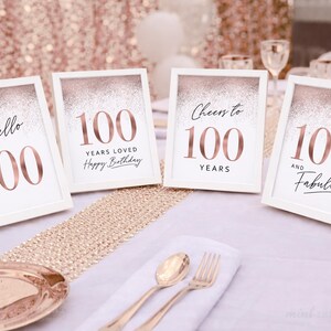 May include: Four white frames containing signs for a 100th birthday celebration. The signs feature rose gold glitter accents and text including Hello 100, 100 Years Loved Happy Birthday, Cheers to 100 Years, and 100 and Fabulous in rose gold and black.