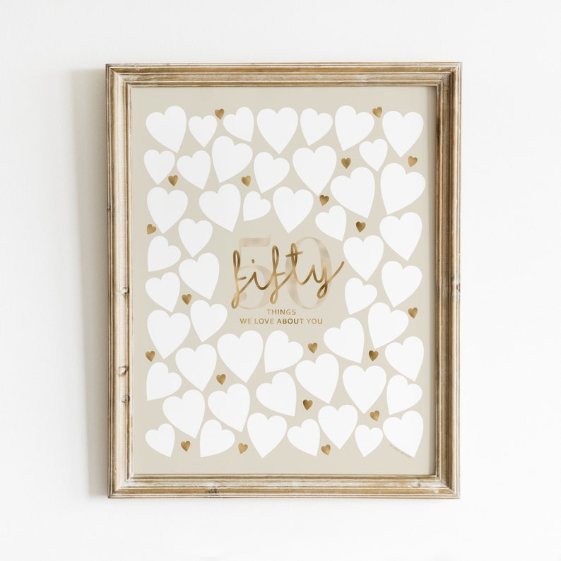 A beige poster with a gold frame. The poster has a gold inscription that says "fifty things we love about you". There are 50 white hearts on the poster.