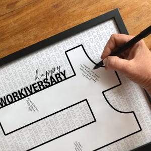 Può includere: Un cartello bianco incorniciato con un bordo nero a forma di numero 11. Lo sfondo presenta un testo ripetuto che recita CONGRATULATIONS ON 11 YEARS OF SERVICE. Il cartello include il testo HAPPY WORKIVERSARY e note scritte a mano.