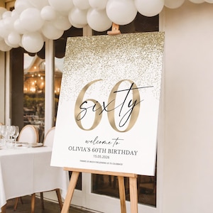 May include: A white sign on a wooden easel with gold glitter at the top and gold numbers that read "60". The word "sixty" is written in a cursive font. Below the numbers is the text "welcome to OLIVIA'S 60TH BIRTHDAY 15.05.2026".