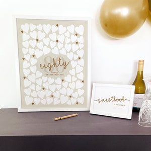 May include: A large white-framed poster featuring many white hearts and the gold text eighty THINGS WE LOVE ABOUT YOU, alongside a smaller white-framed sign with the gold text guestbook PLEASE SIGN and a thin gold pen on a dark surface.