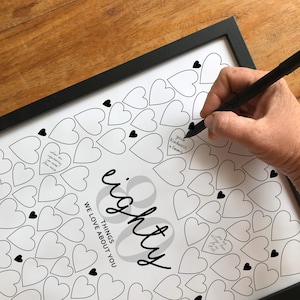 May include: A framed white poster featuring the black text eighty THINGS WE LOVE ABOUT YOU surrounded by numerous outlined hearts. A black pen is writing inside one heart, with handwritten notes visible in three other hearts on the page.